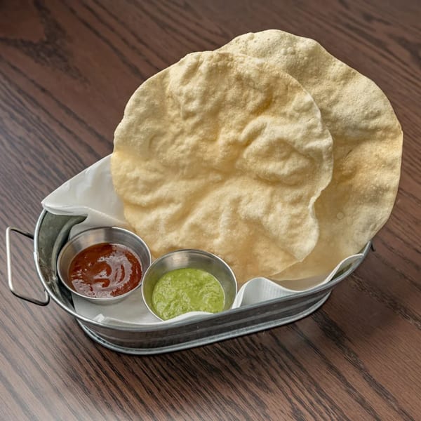 Crispy Indian papadums served in a metal basket with red and green dipping sauces on a wooden table.